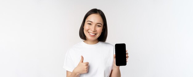 Portret pięknej azjatyckiej kobiety pokazującej kciuk w górę i uśmiechający się ekran smartfona podczas reklamowania aplikacji telefonu komórkowego na białym tle