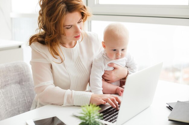 Portret pięknej biznesowej kobiety siedzącej przy stole i pracującej na laptopie, trzymając swoje małe dziecko