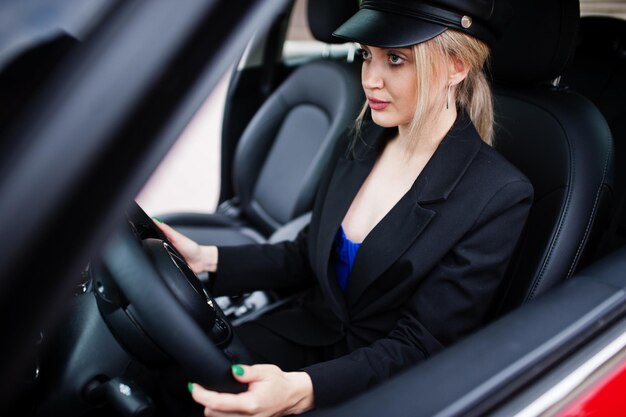 Portret pięknej blond seksownej kobiety modelki w czapce i całej czerni z jasnym makijażem siedzieć i prowadzić czerwony miejski samochód