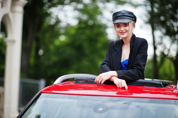 Bezpłatne zdjęcie portret pięknej blond seksownej kobiety modelki w czapce i całej czerni z jasnym makijażem w czerwonym samochodzie miejskim z otwartym szyberdachem