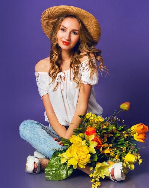 Portret pięknej blondynki młodej damy w stylowym słomkowym kapeluszu letnim trzymając kolorowy wiosenny bukiet kwiatów w pobliżu fioletowego tła ściany.
