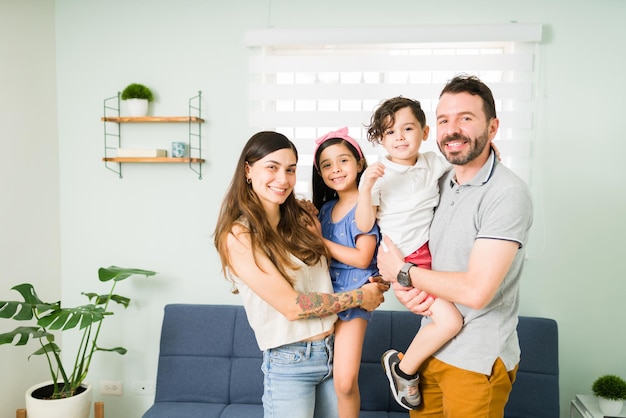 Portret pięknej czteroosobowej rodziny z uroczymi małymi dziećmi uśmiechającymi się i nawiązującymi kontakt wzrokowy podczas wspólnego relaksującego dnia w domu