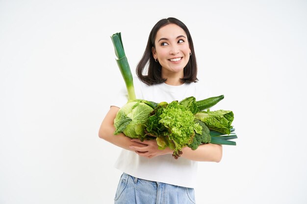 Portret pięknej i zdrowej azjatyckiej kobiety trzymającej zielone warzywa oraniczne i uśmiechającej się szczęśliwie