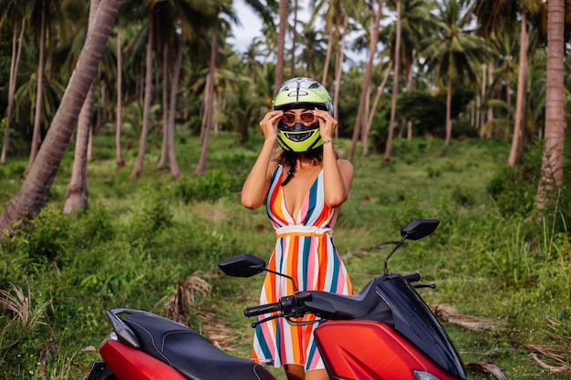 Portret pięknej kobiety jeźdźca w żółto-zielonym kasku motocyklowym i kolorowej lekkiej letniej sukience w dżungli na tropikalnym polu pod palmami.