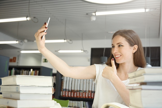 Portret pięknej kobiety trzymającej telefon komórkowy, robiącej selfie przed wnętrzem biblioteki, kobieta wyglądająca na szczęśliwą, uśmiechnięta i pozująca z kciukami w górę w otoczeniu książek i podręczników