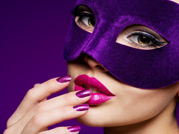 Portret pięknej kobiety z fioletowymi paznokciami i fioletową maską teatralną na twarzy.