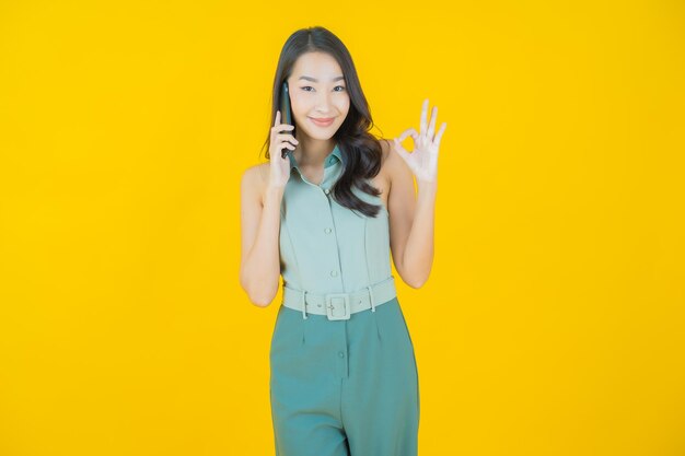 Portret pięknej młodej azjatyckiej kobiety uśmiecha się inteligentnym telefonem komórkowym na żółtej ścianie