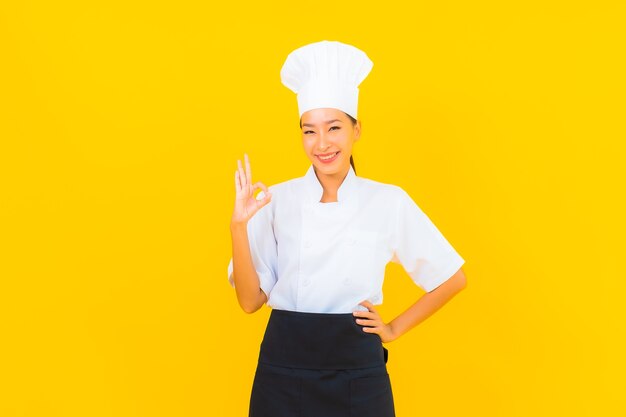 Portret pięknej młodej azjatyckiej kobiety w mundurze szefa kuchni lub kucharza z kapeluszem na żółtym tle na białym tle