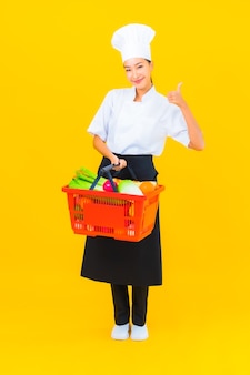 Portret pięknej młodej azjatyckiej kucharki z koszem spożywczym z supermarketu na żółtym tle na białym tle