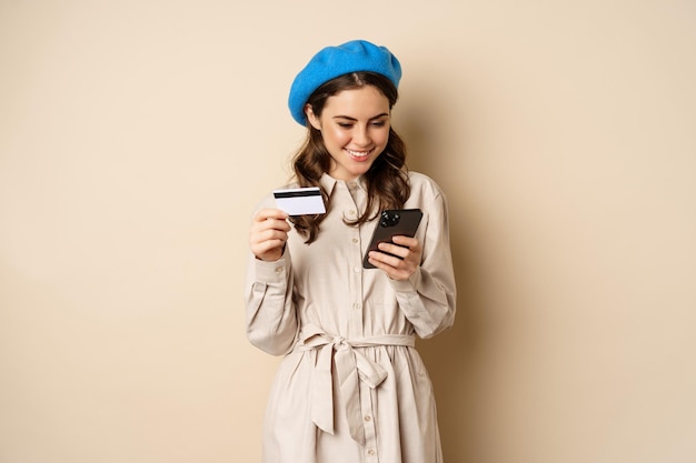 Bezpłatne zdjęcie portret pięknej młodej kobiety, 25 lat, korzystającej z aplikacji do zakupów telefonu komórkowego, karty kredytowej, dostawy zamówienia, zakupu, stojąc na beżowym tle