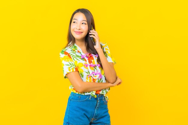 Portret pięknej młodej kobiety azjatyckie na sobie kolorową koszulę za pomocą inteligentnego telefonu komórkowego na żółtej ścianie
