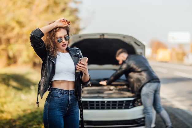 Portret pięknej młodej kobiety za pomocą jej telefonu komórkowego wzywa do pomocy dla samochodu.