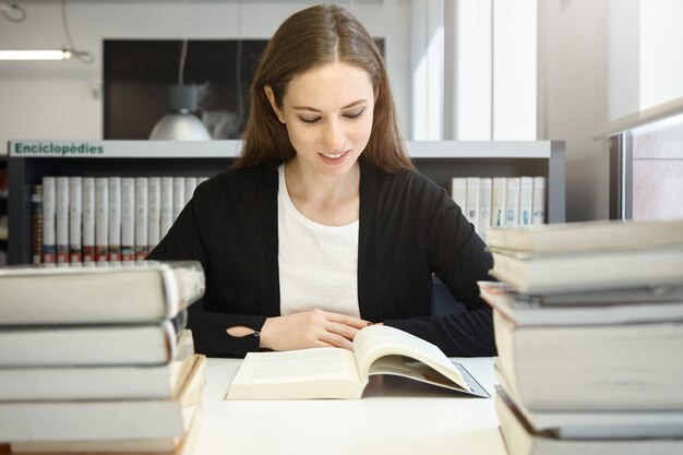 Portret pięknej młodej profesor brunetki w czarnej kurtce, czytanie instrukcji lub podręcznika, uśmiechnięta, przygotowująca się do wykładu na uniwersytecie, siedząca w bibliotece przed stosami książek