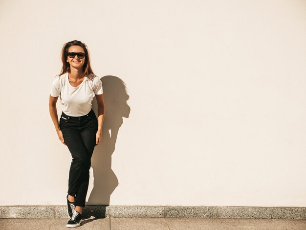 Portret pięknej modelki w okularach przeciwsłonecznych. Kobieta ubrana w letni hipster biały T-shirt i dżinsy. Modna kobieta pozuje przy ścianie na ulicy