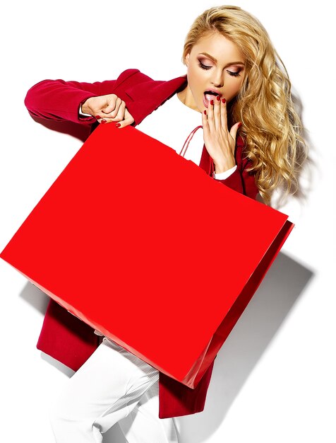 portret pięknej ślicznej szczęśliwej słodkiej zdziwionej blondynki kobiety dziewczyny mienia w jej rękach duża torba na zakupy w modniś czerwieni ubraniach odizolowywających na bielu