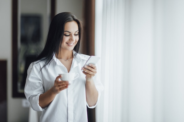 Portret pięknej szczęśliwej dziewczyny brunetka rano z kawą i telefonem w pobliżu okna.