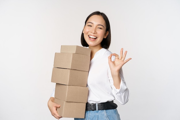 Portret pięknej szczęśliwej kobiety niosącej pudła z zamówieniami Bizneswoman z dostawą niosącą i pokazujący w porządku znak białe tło
