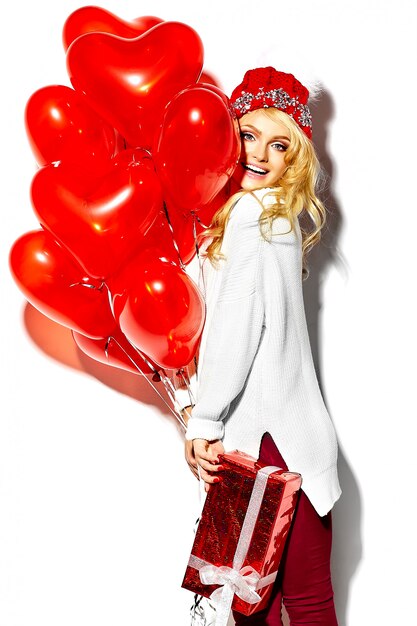 portret pięknej szczęśliwej słodkiej uśmiechniętej blondynki kobiety dziewczyny trzymającej w rękach duże pudełko na prezenty świąteczne i balony serca w dorywczo czerwonych ubraniach zimowych hipster, w białym ciepłym swetrze