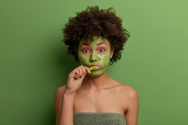Portret pięknej uroczej Afroamerykanki ma poranne rutynowe zabiegi, nosi maseczkę przeciwstarzeniową na twarz dla odmłodzenia, myje zęby, zawinięta w ręcznik kąpielowy, odizolowana na zielonej ścianie.