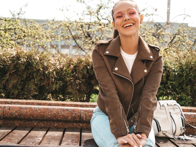 Portret Pięknej Uśmiechniętej Modelki Brunetka Ubrana W Letnie Ubrania Hipster I Dżinsy Modna Dziewczyna Siedzi Na ławce Na Ulicy Zabawna I Pozytywna Kobieta W Okularach Przeciwsłonecznych