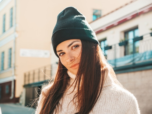 Portret piękny uśmiechający się model. Kobieta ubrana w ciepły, hipsterski biały sweter i czapkę. Pozuje na ulicy