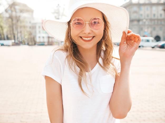 Portret piękny uśmiechnięty blondynu model ubierał w lato modnisiu odziewa. Modna dziewczyna pozuje na ulicy w okrągłe okulary przeciwsłoneczne. Śmieszna i pozytywna kobieta ma zabawę w kapeluszu