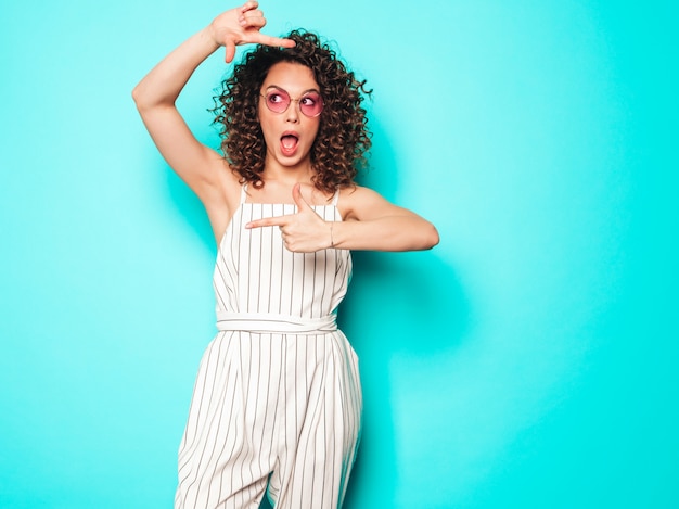 Portret Piękny Uśmiechnięty Model Z Fryzurą Afro Loki Ubrany W Letnie Ubrania Hipster. Seksowna Beztroska Dziewczyna Pozuje W Pobliżu Niebieską ścianą. Modna Kobieta Sprawia, że Ramka Na Zdjęcia Przez Ręce