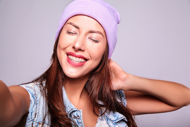 Portret piękny uśmiechnięty śliczny brunetki kobiety model w przypadkowych letnich cajgach odziewa bez makeup w purpurowej czapie robi selfie fotografii na telefonie odizolowywającym na szarość