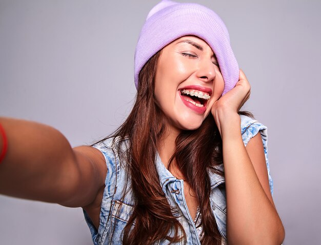 Portret piękny uśmiechnięty śliczny brunetki kobiety model w przypadkowych letnich cajgach odziewa bez makeup w purpurowej czapie robi selfie fotografii na telefonie odizolowywającym na szarość