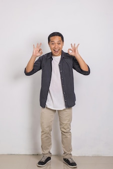 Portret podekscytowanego przystojnego azjatyckiego mężczyzny w koszuli uśmiechającego się i pokazującego znak ok z dwiema rękami w aparacie