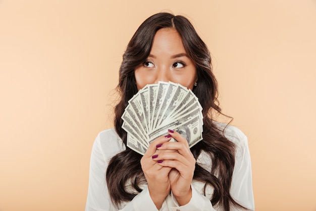 Portret pomyślnej azjatykciej kobiety nakrywkowy usta z fan 100 dolarowych rachunków satysfakcjonuje o pensi lub dochodzie pozuje nad beżowym tłem