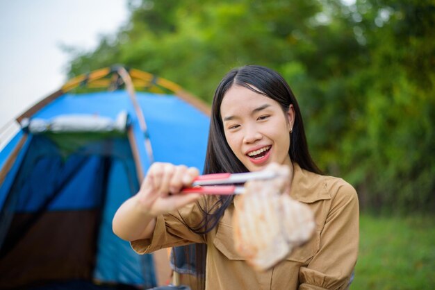 Portret portret szczęśliwej młodej azjatyckiej kobiety kempingowej samotnej grillowanej wieprzowiny na patelni piknikowej i gotowania jedzenia siedząc na krześle na kempingu