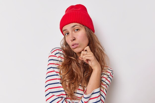 Portret poważna pewna siebie młoda kobieta trzyma rękę pod brodą uważnie patrzy na kamerę nosi czerwony kapelusz i swobodny sweter w paski na białym tle nad białym tłem, uważa coś uważnie