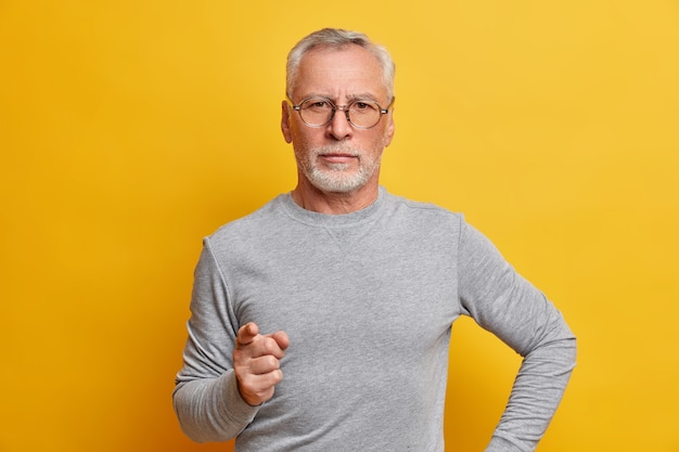 Portret poważnego, surowego brodatego mężczyzny ostrzega palcem, który udziela mądrej rady, nosi szary sweter z długimi rękawami, wygląda pewnie z przodu odizolowany na żółtej ścianie