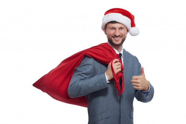 Portret pozytywnego Świętego Mikołaja w szarym garniturze, czerwonej czapce i pełnej torbie na ramieniu, uśmiechnięty i pokazujący super.