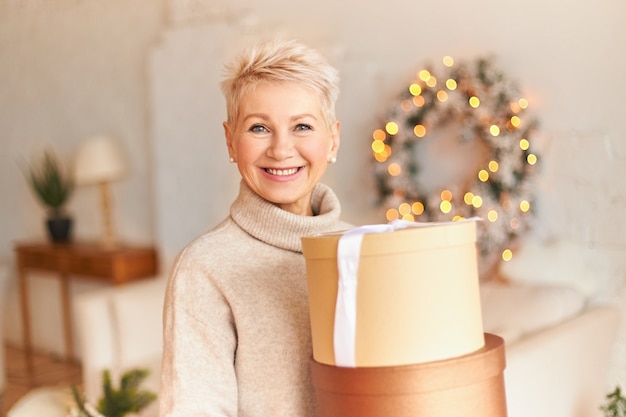 Bezpłatne zdjęcie portret pozytywnej dojrzałej kobiety w swetrze z promiennym szczęśliwym uśmiechem pozuje w przytulnym salonie z świątecznymi dekoracjami, trzymając pudełko z prezentami od syna. wesołych świąt
