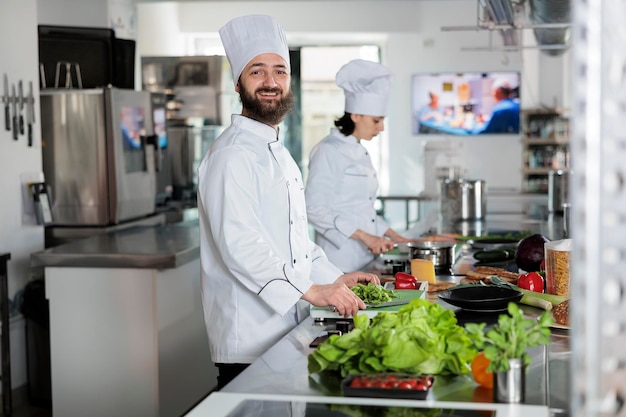 Portret pozytywny uśmiechający się szef kuchni stojący w profesjonalnej kuchni restauracji, patrząc na kamery. Przekonany, człowiek ubrany w biały mundur przemysłu spożywczego podczas przygotowywania pysznego posiłku.