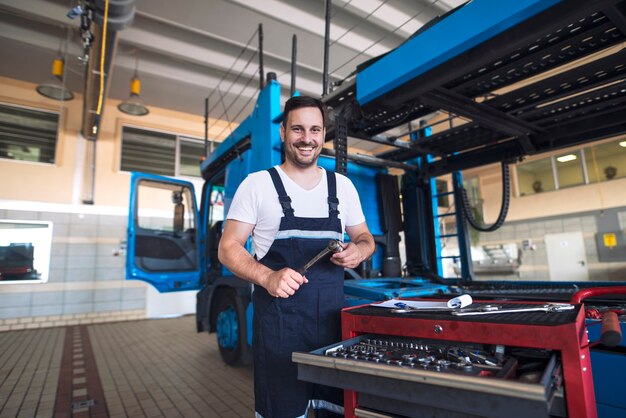 Portret pozytywny uśmiechnięty serwisant ciężarówki z narzędziami stojący przy pojeździe ciężarówki w warsztacie