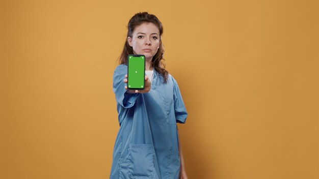 Bezpłatne zdjęcie portret profesjonalnego medyka, trzymając smartfon i wysyłając sms-y i pokazując projekt makiety zielonego ekranu w aparacie w studio. lekarz w mundurze szpitalnym za pomocą urządzenia z ekranem dotykowym, prezentując miejsce na kopię.