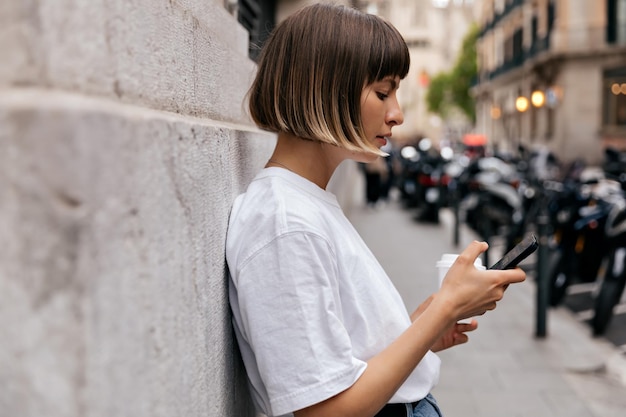 Portret profil stylowa dziewczyna krótkowłosa w biały tshirt przewijanie smartfona w mieście Modna dziewczyna kaukaski ze smartfonem pozowanie odkryty z uśmiechem