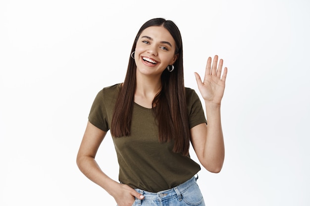 Portret przyjaznej młodej szczęśliwej kobiety macha ręką, aby się przywitać, witając cię gestem powitania, żegnając się, stojąc nad białą ścianą