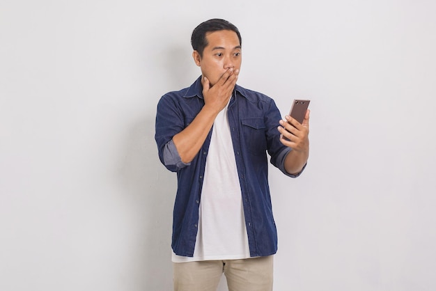 Portret przypadkowego azjatyckiego mężczyzny jest zszokowany i zaniemówił, patrząc na telefon komórkowy