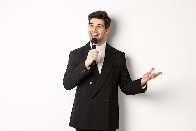 Portret przystojnego mężczyzny w czarnym garniturze śpiewającego piosenkę, trzymającego mikrofon i przemawiającego, stojącego na białym tle