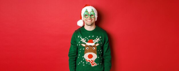 Portret przystojnego mężczyzny w zielonym świątecznym swetrze santa hat i imprezowych okularach uśmiechający się szczęśliwy standi