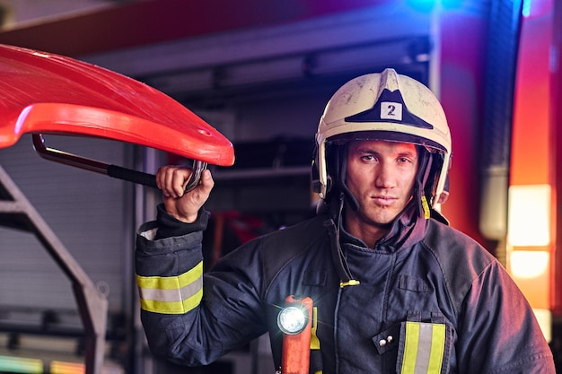 Portret przystojnego strażaka w mundurze ochronnym z latarką, w tym stojący w garażu straży pożarnej i patrzący na kamerę