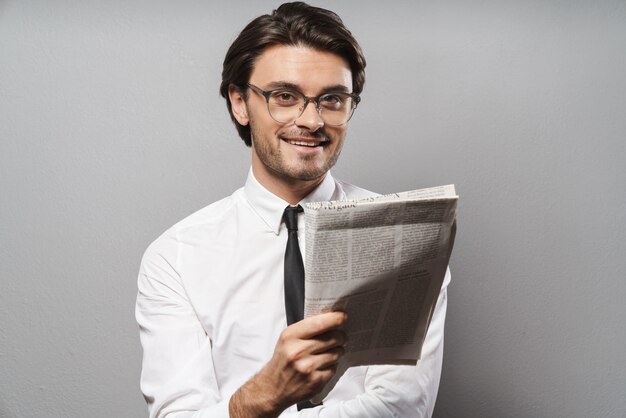 Portret przystojnego uśmiechniętego młodego biznesmena w garniturze stojącego na białym tle nad szarą ścianą, czytającego gazetę