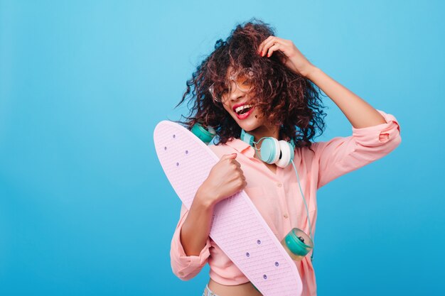 Portret przystojny afrykański modelka w słuchawkach trzyma nową deskorolkę i uśmiecha się. Roześmiana mulatka w modnej różowej koszuli bawi się kręconymi brązowymi włosami.