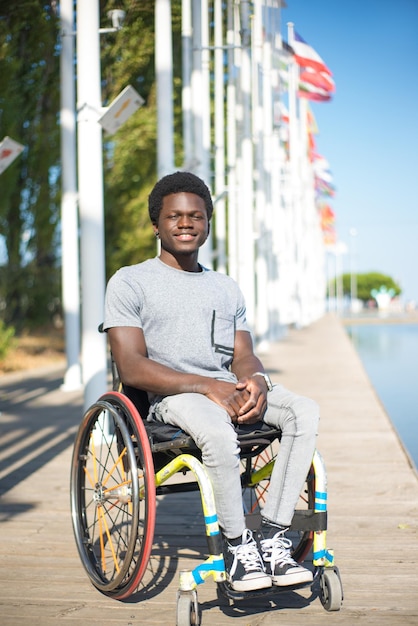 Portret przystojny mężczyzna na wózku inwalidzkim. African American cz?owiek w dorywczo ubrania na nasyp, u?miechaj?c si?. Błękitne niebo i flagi w tle. Portret, piękno, koncepcja szczęścia