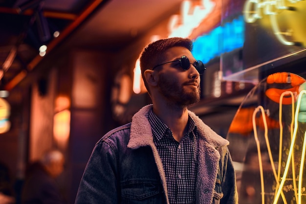 Portret przystojny mężczyzna ubrany w dżinsowy płaszcz i okulary przeciwsłoneczne, stojący w nocy na ulicy. Podświetlane szyldy, neony, światła.
