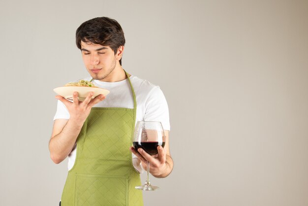 Portret przystojny mężczyzna w fartuchu trzyma talerz z makaronem i kieliszek wina.